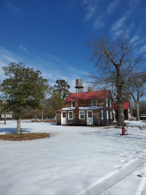 A rustic wooden house with a red roof and water tower, surrounded by snow and trees under a clear blue sky.