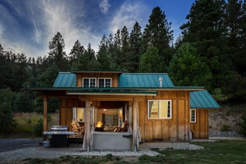 A cozy wooden cabin with a green metal roof, surrounded by trees, featuring a porch and a hot tub.