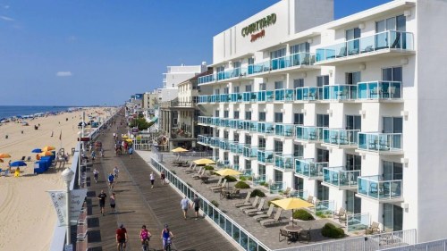 A beachfront hotel with balconies, overlooking a boardwalk filled with people and umbrellas on the sandy beach.