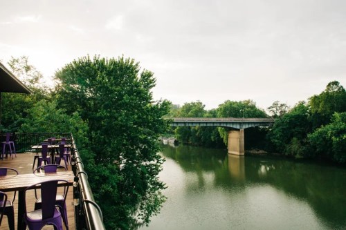 A serene river view with a bridge in the background, surrounded by lush greenery and a deck with purple chairs.