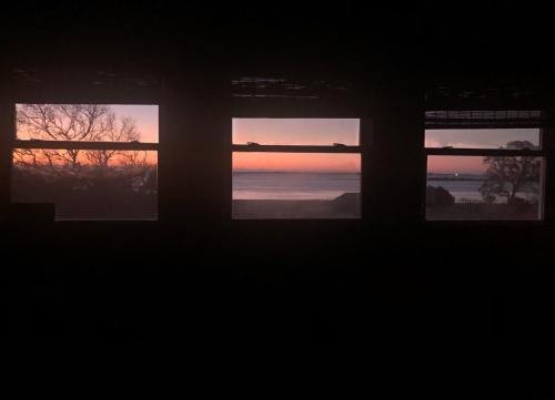Three windows frame a serene sunset over the ocean, with silhouettes of trees against a colorful sky.