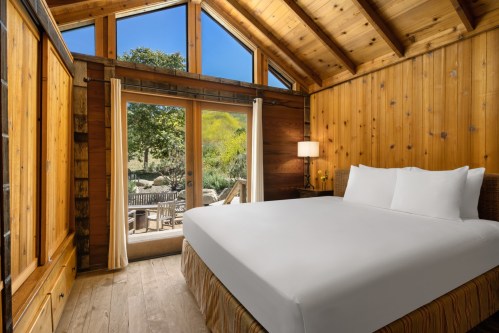 Cozy wooden bedroom with a large bed, large windows, and a view of a patio and greenery outside.