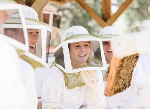 A group of people in beekeeping suits smiling and observing a honeycomb frame outdoors.