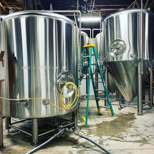 Two large stainless steel fermentation tanks in a brewery, with a ladder and hoses on the floor.