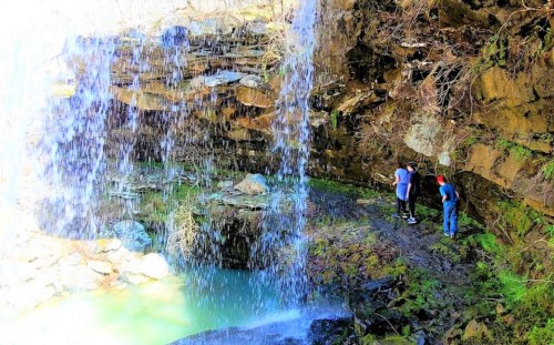 Two people stand near a waterfall, surrounded by rocky cliffs and a pool of water below. Sunlight filters through.