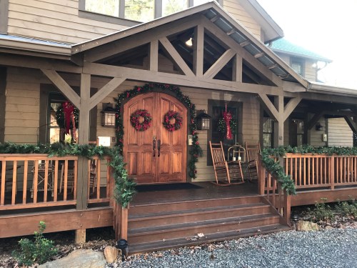 A cozy wooden house entrance decorated with wreaths and garlands for the holidays, featuring rocking chairs on the porch.