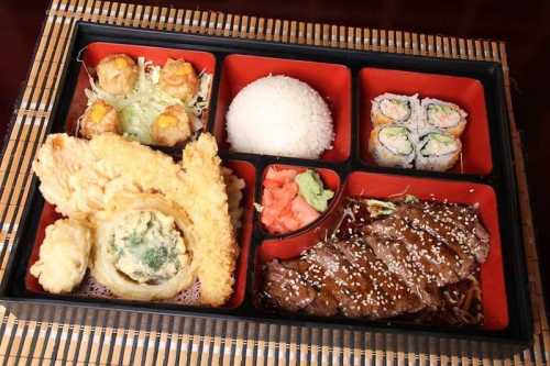 A colorful bento box featuring tempura, rice, sushi rolls, and grilled meat with vegetables.