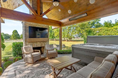 Cozy outdoor space with a hot tub, seating area, and a stone fireplace under a wooden pergola. Lush greenery in the background.