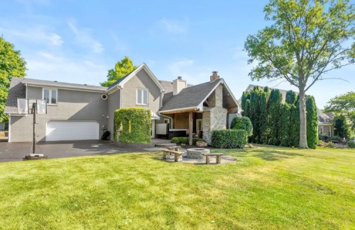 A modern house with a stone entrance, surrounded by greenery and a lawn, featuring a fire pit and basketball hoop.