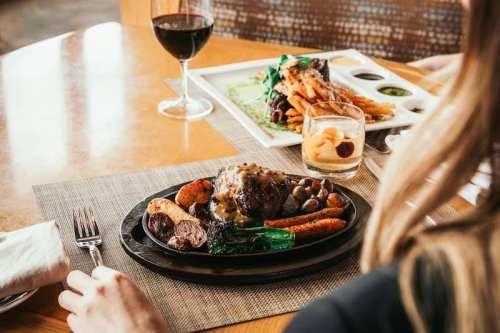 A person sits at a table, enjoying a plate of steak with vegetables, alongside a glass of wine and another dish in the background.