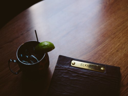 A cocktail with a lime wedge sits next to a leather menu on a wooden table.