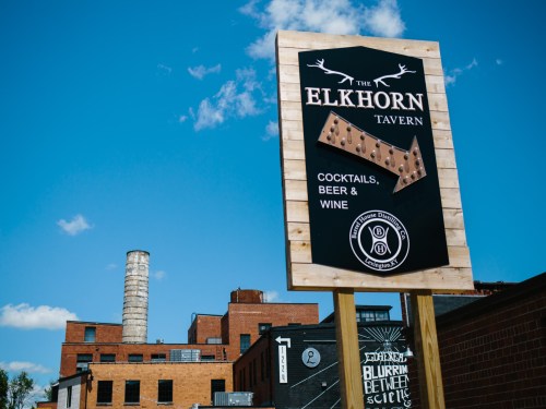 Sign for The Elkhorn Tavern featuring cocktails, beer, and wine, set against a blue sky and urban backdrop.