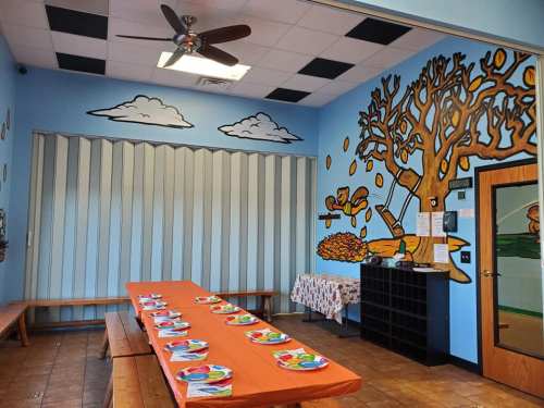 A colorful dining area with a long orange table set for a meal, featuring a mural of a tree and clouds on the walls.