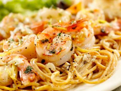 A close-up of shrimp pasta with herbs and spices, garnished with lemon and served with a side of greens.