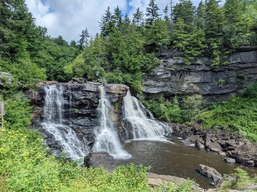 A scenic waterfall cascading over rocky cliffs, surrounded by lush greenery and trees under a partly cloudy sky.