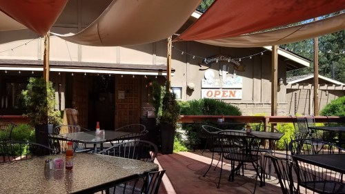 Outdoor dining area with tables and chairs, shaded by fabric canopies, in front of a restaurant with an "OPEN" sign.