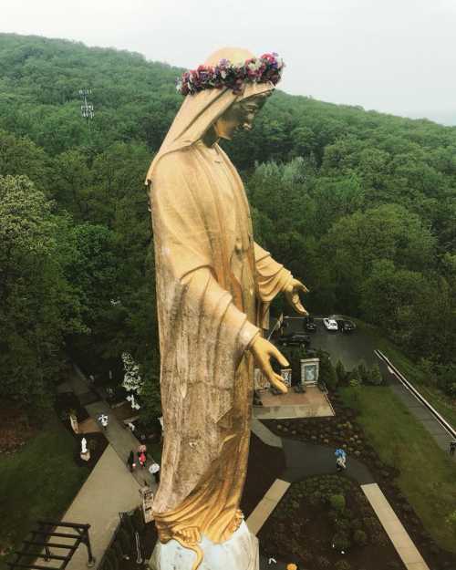 A large golden statue of a figure with a floral crown, surrounded by lush green trees and visitors below.