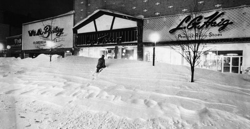 A person trudges through deep snow in front of closed storefronts at night, illuminated by streetlights.