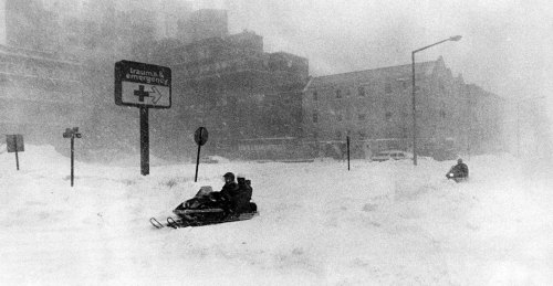 Two snowmobiles navigate through a snowy, blizzard-like scene near a hospital sign, with buildings partially obscured by snow.