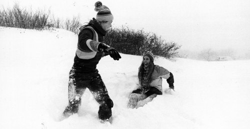 Two people play in deep snow, one laughing while the other runs, creating a joyful winter scene.