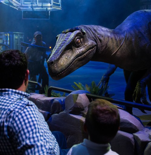 A lifelike dinosaur animatronic interacts with an audience in a dimly lit arena.