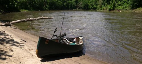 A canoe rests on a sandy riverbank, with trees lining the water's edge and a fishing rod visible inside the canoe.