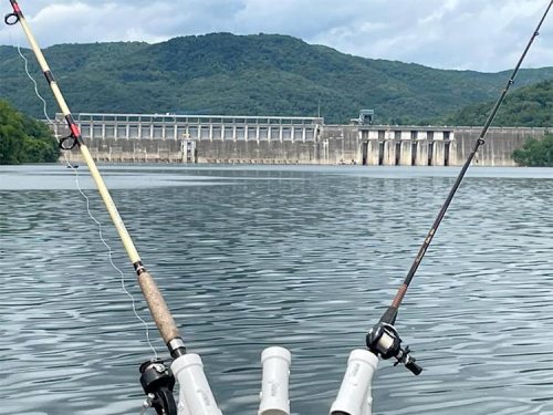 Two fishing rods are positioned over calm water, with a dam and mountains in the background under a cloudy sky.
