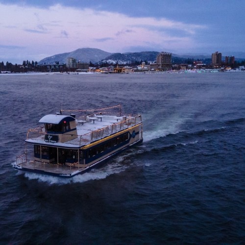 A decorated ferry boat sails on a calm waterway with a snowy mountain and city skyline in the background.