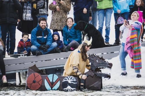 A crowd watches as a person in a Viking costume sits by a snowy waterway, surrounded by children and a dog.