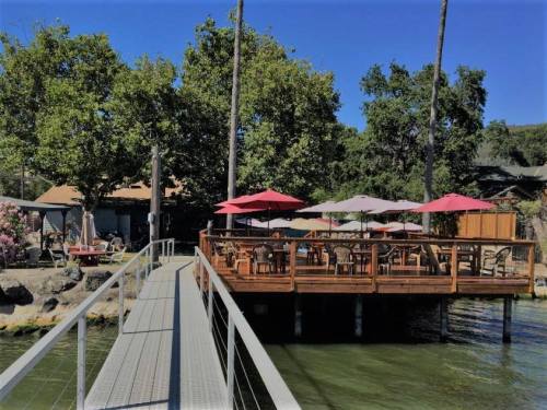 A wooden deck with umbrellas overlooks a calm waterway, connected by a walkway, surrounded by trees and a cozy building.