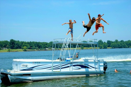 Three boys jump off a pontoon boat into a lake on a sunny day, with trees lining the shore in the background.