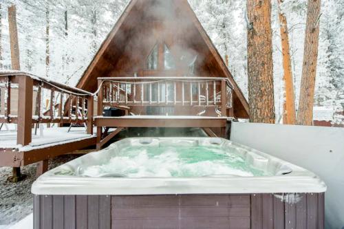 A hot tub with bubbling water sits outside a cozy cabin surrounded by snow-covered trees. Steam rises in the cold air.