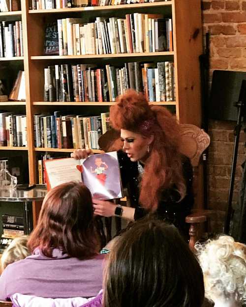 A performer in a sparkly outfit reads a children's book to an audience in a cozy library setting.