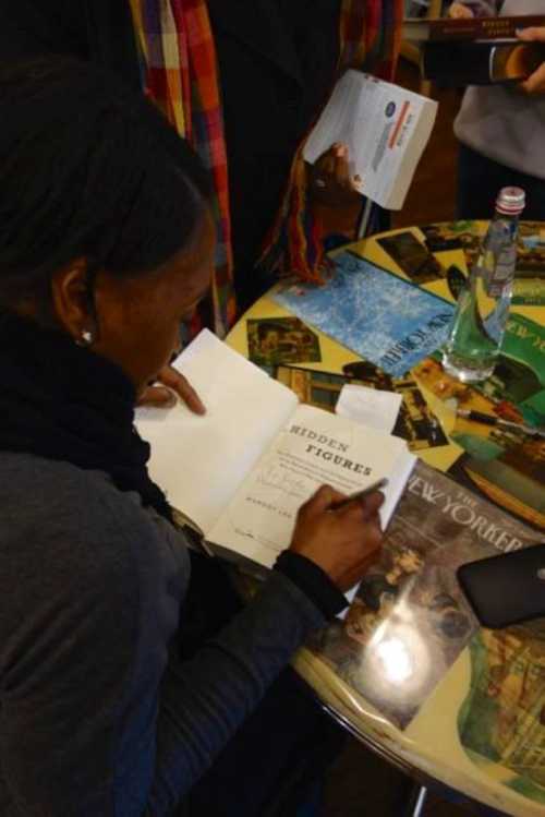 A person signing a book titled "Hidden Figures" at a table covered with magazines and a water bottle.