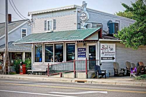 A quaint restaurant with a green metal roof, large windows, and outdoor seating, located on a street.