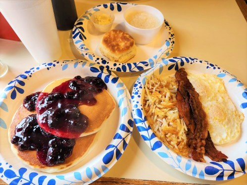 A plate of pancakes with berry syrup, a side of hash browns, bacon, and an egg, alongside a biscuit and grits.