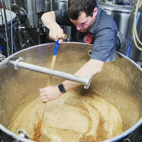 A brewer stirs a large vat of bubbling liquid in a brewery, surrounded by stainless steel equipment.