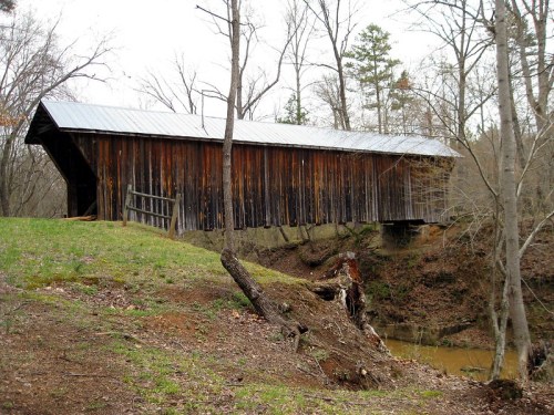 A rustic covered bridge spans a small creek, surrounded by bare trees and grassy banks in a serene landscape.