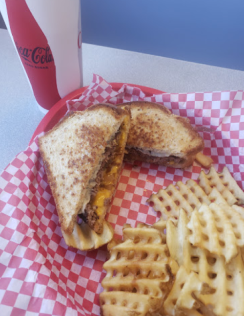 A toasted sandwich with melted cheese and a side of waffle fries, served in a red-checkered basket with a soda.
