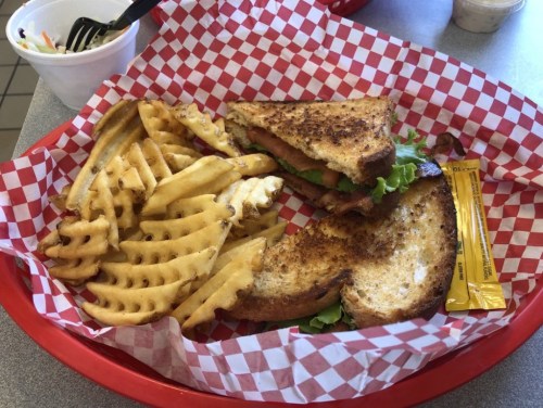 A red basket with a grilled sandwich, lettuce, and waffle fries, accompanied by a packet of sauce.