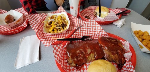 A table with red-checkered trays featuring ribs, a burger, fries, and a drink, with napkins and utensils.