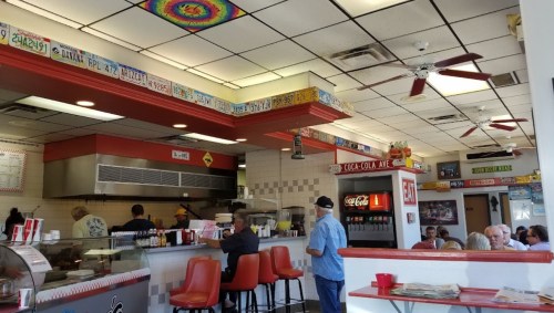 A colorful diner interior with red seating, a counter, and patrons enjoying their meals. Ceiling fans and decor visible.