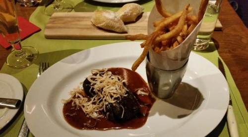 A plate of meat in sauce topped with cheese, accompanied by a glass of fries and a bread roll on a colorful table setting.