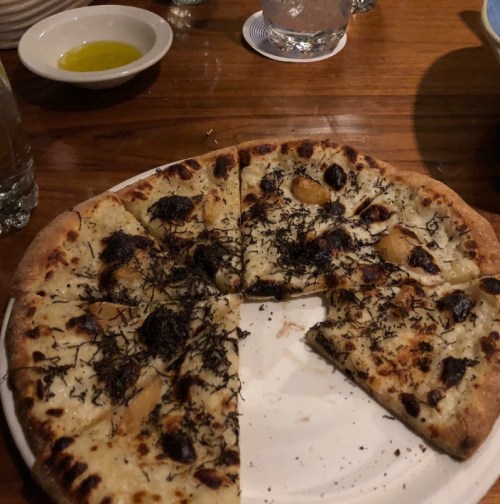 A pizza with a thin crust topped with cheese, black truffles, and sliced mushrooms, partially sliced on a white plate.