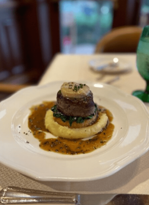 A gourmet dish featuring a tower of meat atop creamy mashed potatoes, garnished with sauce and herbs.