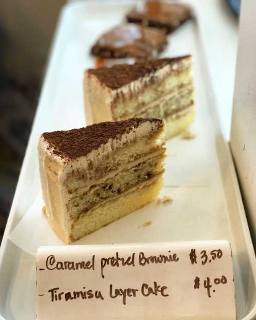 A display of desserts featuring caramel pretzel brownie and tiramisu layer cake with prices on a white tray.