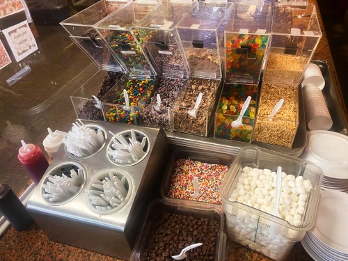 A variety of toppings in clear containers, including candies, sprinkles, and chocolate, with spoons and sauces nearby.