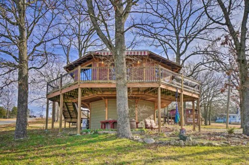 A wooden, elevated house surrounded by trees, featuring a wraparound deck and stairs leading to the ground.
