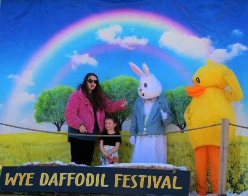 A woman in a pink jacket, a girl in a dress, and costumed characters of a bunny and a duck pose in front of a rainbow backdrop.