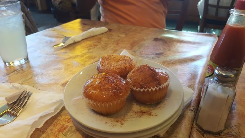 Three golden muffins dusted with cinnamon sugar on a white plate, set on a table with utensils and a drink in the background.